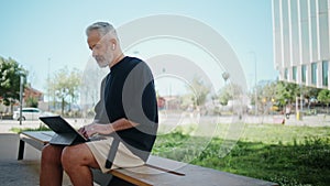 Stylish senior typing computer in urban park. Thoughtful man resting outdoors