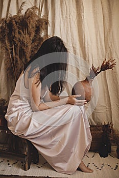 Stylish sensual woman in silk dress holding vase with flowers in bohemian room with dry grass