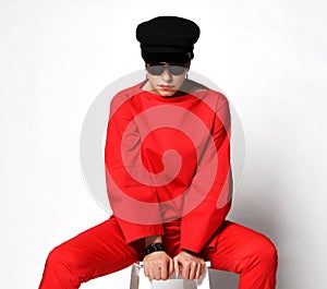 Stylish short haired brunette woman in haute couture casual red pantsuit, sunglasses and black cap sits with legs spread