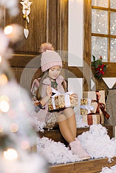 A stylish six-year-old girl in a beret holds a Christmas gift and opens it, sitting on the porch