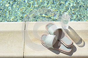 Stylish slippers and glass of water near outdoor swimming pool on sunny day, space for text