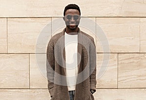 Stylish smiling african man wearing brown knitted cardigan and sunglasses on city street over brick wall