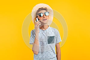 Stylish smiling teen guy in a summer hat, talking on a mobile phone on an yellow background