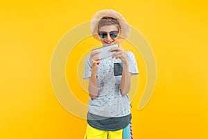 Stylish smiling teen guy in a summer hat using a mobile phone standing on an yellow background