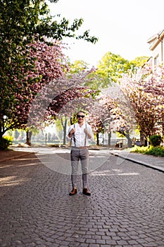 Stylish smiling young man wearing sunglasses, classic white shirt, grey pants and suspenders is standing at city street