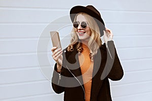 Stylish smiling young woman taking selfie picture by smartphone or video calling wearing black coat and round hat on city street