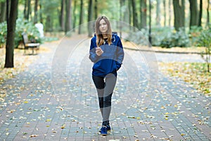 Stylish sporty brunette woman in trendy urban outwear posing at forest city park on cold rainy fall day waiting for a call on