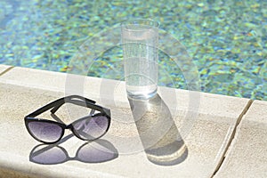 Stylish sunglasses and glass of water near outdoor swimming pool on sunny day