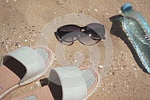 Stylish sunglasses, slippers and bottle of water on sand