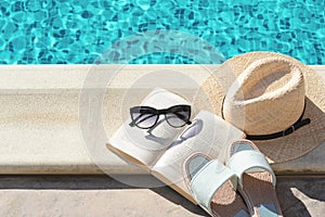 Stylish sunglasses, slippers, straw hat and book at poolside on sunny day. Beach accessories