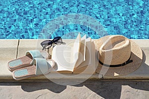 Stylish sunglasses, slippers, straw hat and book at poolside on sunny day. Beach accessories