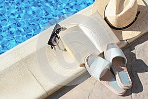 Stylish sunglasses, slippers, straw hat and book at poolside on sunny day. Beach accessories