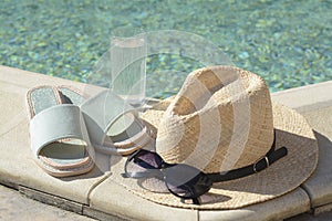 Stylish sunglasses, slippers, straw hat and glass of water at poolside on sunny day. Beach accessories