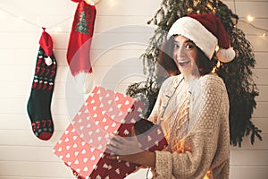 Stylish surprised girl in santa hat opening gift box in christmas decorated room. Young woman in festive cozy sweater opening