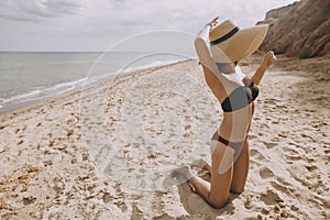 Stylish tan girl in hat sitting on beach. Fashionable young woman covering with straw hat, relaxing on sandy beach near sea.