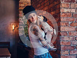 A stylish tattoed blonde female in t-shirt and jeans holds a cute dog.