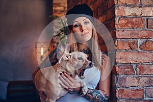 A stylish tattoed blonde female in t-shirt and jeans holds a cute dog.