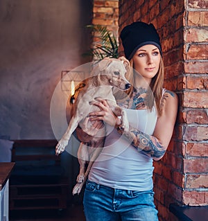 A stylish tattoed blonde female in t-shirt and jeans holds a cute dog.