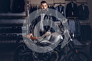 Stylish tattooed bearded man with dressed in black leather jacket and bow tie posing near retro sports motorbike at men