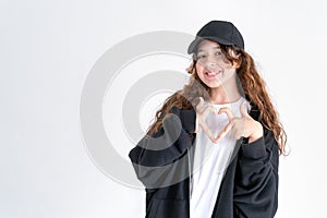 Stylish teen girl with long curly hair in a black hoodie and a black cap shows a heart shape with her fingers and smiles on a