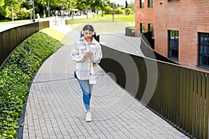 Stylish teen lady in wireless headphones texting on smartphone while walking in the urban city area or college campus