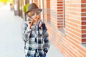 Stylish teenage boy with mobile phone. Hipster kid in sunglasses and modern hat at city street. Young boy talking on the