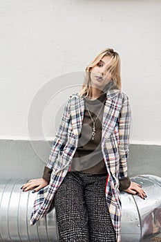Stylish teenager girl with blond hair in oversized gray trendy plaid jacket in vintage trousers sits on silver metal pipe near