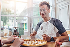 Stylish teenager wearing napkin holding knife and fork ready to eat pizza sitting against window at restaurant