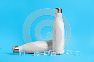 Stylish thermo bottles with water drops and ice cubes on light blue background, space for text