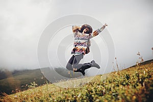 Stylish traveler girl in hat with backpack jumping in mountains.