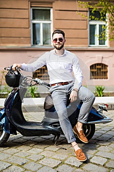 Stylish trendy man wearing modern sunglasses and a formal suit sitting waiting on a motorcycle on city street