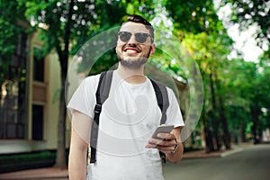 Stylish unshaven hipster wearing white tshirt, black bagpack and sunglasses using a smart phone outdoor