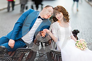 Stylish wedding couple bride in white dress and elegant groom sitting on a bench holding hands