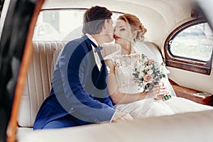 Stylish wedding couple sitting inside of luxury car on white leather, embracing and kissing. elegant groom and bride romantic