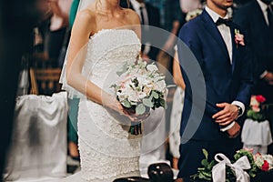 Stylish wedding couple standing in church for holy matrimony. Beautiful bride and groom praying at wooden altar in church, wedding