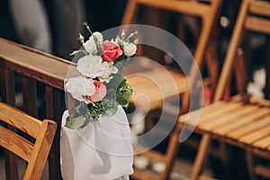 Stylish wedding decor of wooden benches in church for holy matrimony. Beautiful roses and tulle bouquets on wooden chairs,