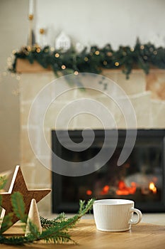 Stylish white cup, wooden star decor on table against stylish festive christmas tree with golden lights and cozy fireplace. Winter