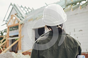 Stylish woman architect with blueprints  looking at unfinished modern farmhouse at construction site. Young female engineer or