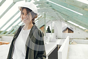 Stylish woman architect or engineer smiling against wooden roof framing of modern farmhouse. Portrait of young happy female in