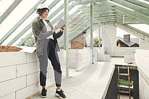 Stylish woman architect or engineer with tablet checking blueprints against wooden roof framing of modern farmhouse. Young happy