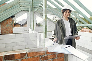 Stylish woman architect or engineer with tablet checking blueprints against wooden roof framing of modern farmhouse. Young happy