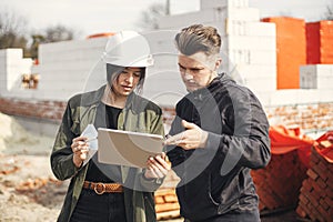 Stylish woman architect with tablet and contractor man checking blueprints at construction site. Young engineer or construction