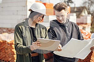 Stylish woman architect with tablet and contractor man checking blueprints at construction site. Young engineer or construction