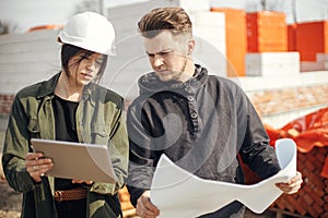 Stylish woman architect with tablet and foreman checking blueprints at construction site. Young engineer and construction workers