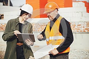 Stylish woman architect with tablet and senior foreman checking blueprints at construction site. Engineer and construction worker