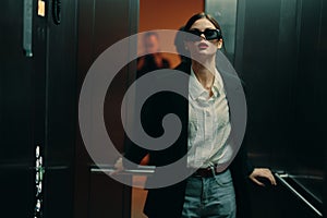 Stylish woman in black jacket and sunglasses posing in elevator, fashion model, dark cinematic light and color, glamor