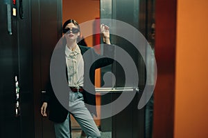 Stylish woman in black jacket and sunglasses posing in elevator, fashion model, dark cinematic light and color, glamor