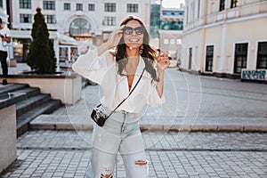 Stylish woman with camera looking up while walking down the street. Outdoor photo of carefree brunette girl in