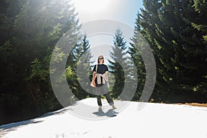 Stylish woman in casual clothes standing in the snow in the woods on a sunny spring day, walking on a mountain resort