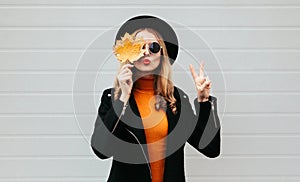 Stylish woman closes her eyes with yellow maple leaves blowing red lips sending sweet air kiss, female model in black coat